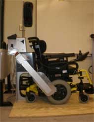 This figure shows three pictures of  a) manual wheelchair secured by 4-point strap-type tiedown; b) top view of UDIG auto docking system;  c) powered wheelchair in rear-facing passenger station (RF-WPS) 